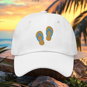 May include: White baseball cap with an embroidered design of two flip-flops. The flip-flops are tan with blue straps. The hat has a curved brim and a button on top. The background shows a beach scene with palm trees.