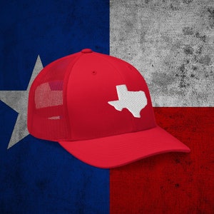May include: Red and white trucker hat with a white outline of the state of Texas on the front. The hat is sitting on a Texas flag.