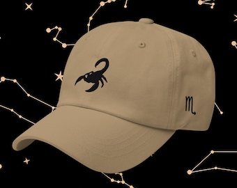 May include: A tan baseball cap with a black embroidered scorpion design on the front. The cap also features the astrological symbol for Scorpio on the side. The background is black with a constellation pattern.