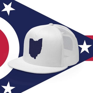 May include: White trucker hat with a navy blue outline of the state of Ohio on the front. The hat is on top of a red, white, and blue flag with a circle in the center.