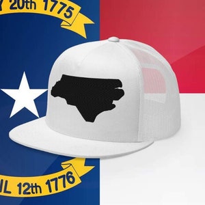 May include: White trucker hat with a black outline of the state of North Carolina on the front. The hat is sitting on a white surface in front of a red, white, and blue flag.