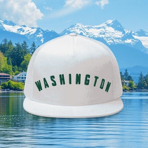 May include: White trucker hat with a green embroidered "WASHINGTON" logo. The hat is photographed against a backdrop of a lake and mountains.
