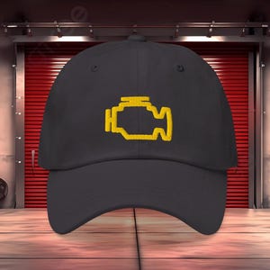 May include: Black baseball cap featuring a yellow engine light symbol. The cap is made of a durable fabric and has a curved brim. A classic, adjustable hat for everyday wear.