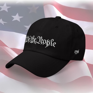 May include: Black baseball cap with the embroidered text "We the People" in white script. The year "1776" is embroidered on the side. The background features an American flag.