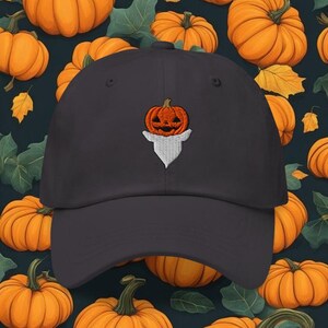 Puede incluir: Gorra de béisbol gris oscuro con un diseño bordado de calabaza naranja. La gorra tiene una visera curva y una construcción clásica de seis paneles. El fondo es un patrón de calabazas naranjas y hojas verdes.