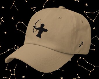 May include: A tan baseball cap featuring a black embroidered Sagittarius symbol, a silhouette of an archer with a bow and arrow. The cap has a curved brim and a small Sagittarius symbol on the side. The background is black with a constellation pattern.