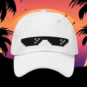 May include: A white baseball cap featuring a pixelated sunglasses design in black. The cap has a curved brim and a classic six-panel construction. The background shows a sunset with palm trees and birds.