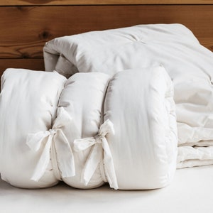May include: A white, quilted, and tied crib bumper with a white, quilted comforter folded on top of it.