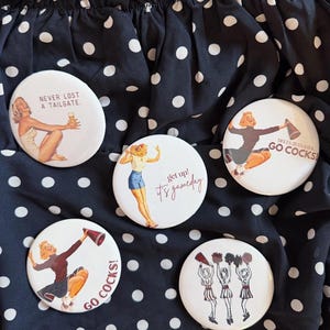May include: Five circular pinback buttons featuring vintage-style illustrations. One shows a person with the text NEVER LOST A TAILGATE. Another shows a person with get up! it's gameday. Two show cheerleaders with GO COCKS! and one shows three cheerleaders.