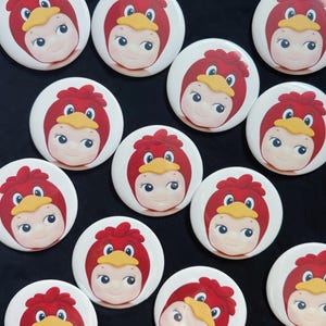 May include: A collection of eleven circular pin-back buttons, each featuring a graphic illustration of a face wearing a red bird-themed hood with a yellow beak. The buttons are arranged in a scattered pattern, showcasing the identical character design.