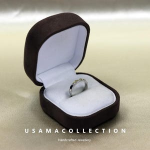 May include: A silver ring with a decorative band, displayed in an open brown velvet jewelry box with a white interior. The ring has small, gold-colored accents. The text "USAMA COLLECTION Handcrafted Jewellery" is at the bottom.