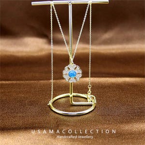 Könnte beinhalten: Eine goldfarbene Halskette mit einem blütenförmigen Anhänger mit einem blauen Mittelstein und klaren Steinen. Die Halskette wird auf einem goldenen Schmuckständer präsentiert. Der Ständer hat eine kreisförmige Basis und eine vertikale Stütze. Der Text "USAMA COLLECTION Handcrafted Jewellery" steht unten.