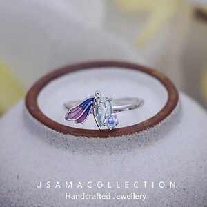 May include: A silver ring featuring a dragonfly design with purple and blue wings, a light blue rectangular gemstone, and a small blue flower. The ring is set against a neutral background. The text "USAMA COLLECTION Handcrafted Jewellery" is visible.