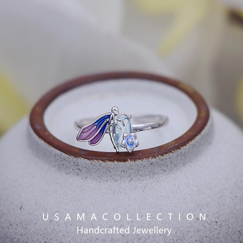 May include: A silver ring featuring a dragonfly design with purple and blue wings, a light blue rectangular gemstone, and a small blue flower. The ring is set against a neutral background. The text "USAMA COLLECTION Handcrafted Jewellery" is visible.