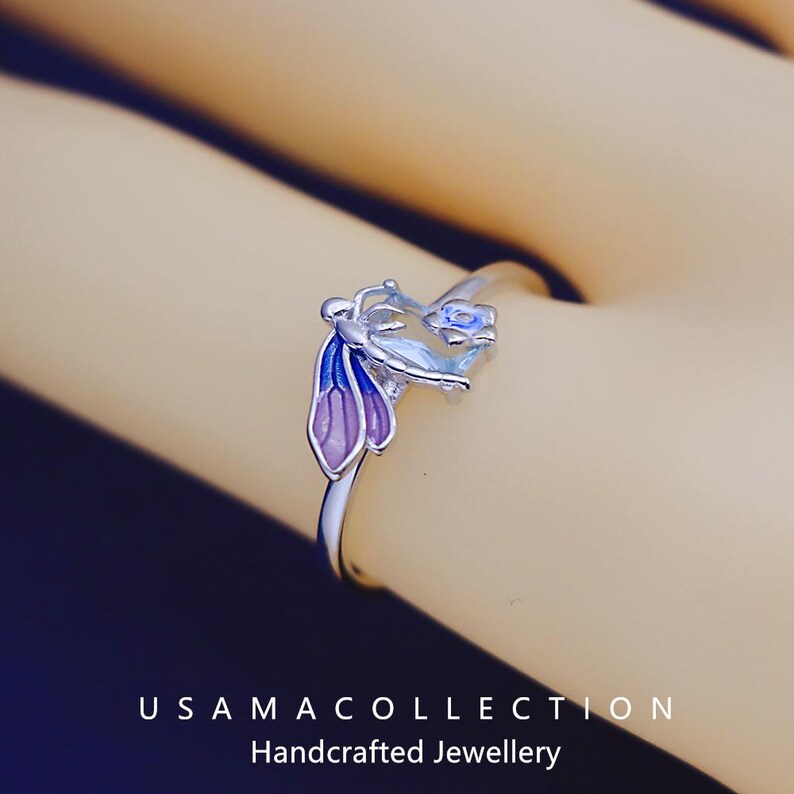 May include: A silver ring with a floral design featuring blue and purple enamel accents. The ring has a delicate band and a detailed flower motif. The text "USAMA COLLECTION Handcrafted Jewellery" is at the bottom of the image.