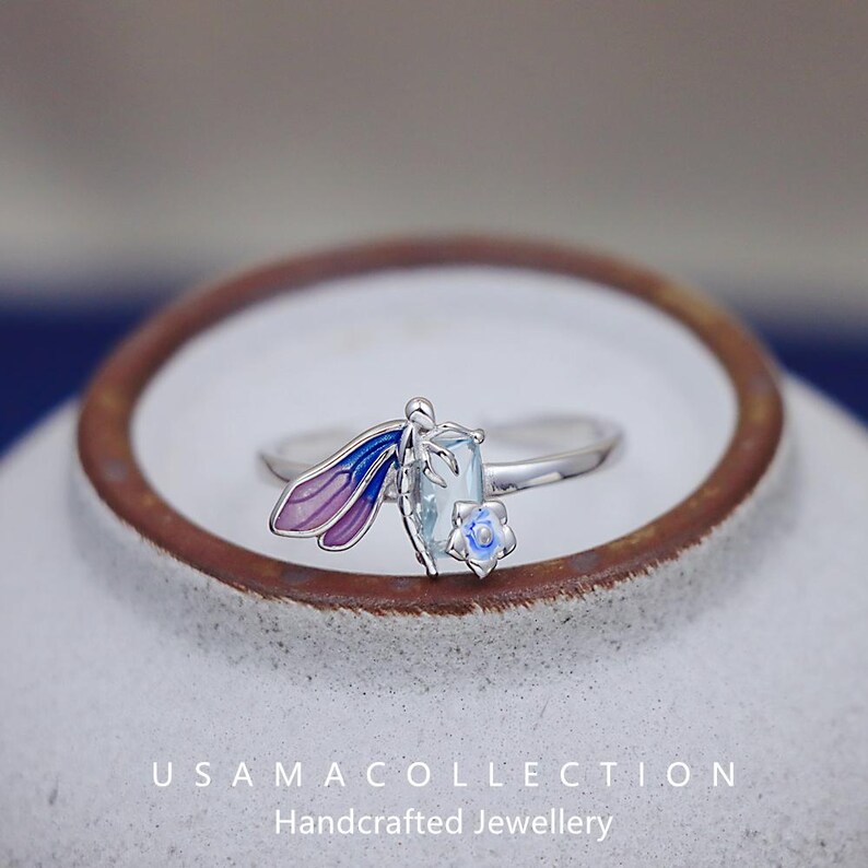May include: A silver ring featuring a dragonfly design with blue and purple wings, a light blue rectangular gemstone, and a small blue flower. The ring is set against a white background with the text "USAMA COLLECTION Handcrafted Jewellery".