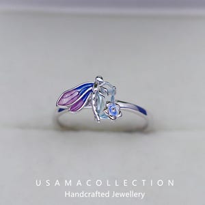 May include: A silver ring featuring a dragonfly design with blue and purple enamel wings. The ring has a light blue gemstone and a small blue flower accent. The text "USAMA COLLECTION Handcrafted Jewellery" is at the bottom.