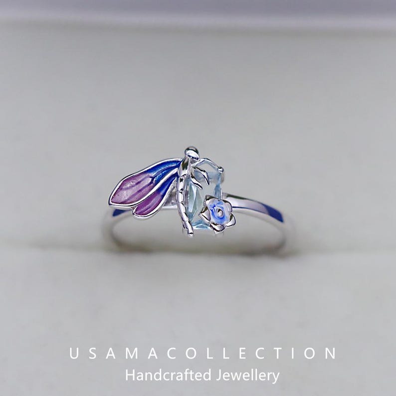 May include: A silver ring featuring a dragonfly design with blue and purple enamel wings. The ring has a light blue gemstone and a small blue flower accent. The text "USAMA COLLECTION Handcrafted Jewellery" is at the bottom.