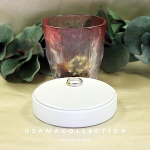 May include: A silver ring with a gold accent sits on a white display stand. The ring is the focal point, with a blurred red and clear glass in the background. The text "USAMA COLLECTION Handcrafted Jewellery" is at the bottom.