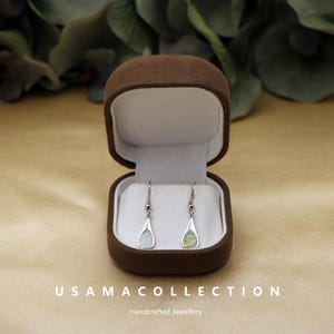 May include: A pair of silver teardrop earrings with iridescent opal inlays displayed in an open brown velvet jewelry box. The earrings hang from silver hooks. The box sits on a gold-colored surface. The text "USAMA COLLECTION" and "Handcrafted Jewellery" are visible.