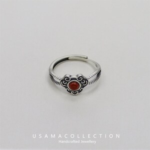 Pode incluir: Um anel prateado com uma banda ajustável. O anel apresenta uma pedra vermelha redonda central, inserida num design decorativo em forma de nuvem. A banda tem um padrão texturizado. O texto "USAMA COLLECTION Handcrafted Jewellery" está na parte inferior.