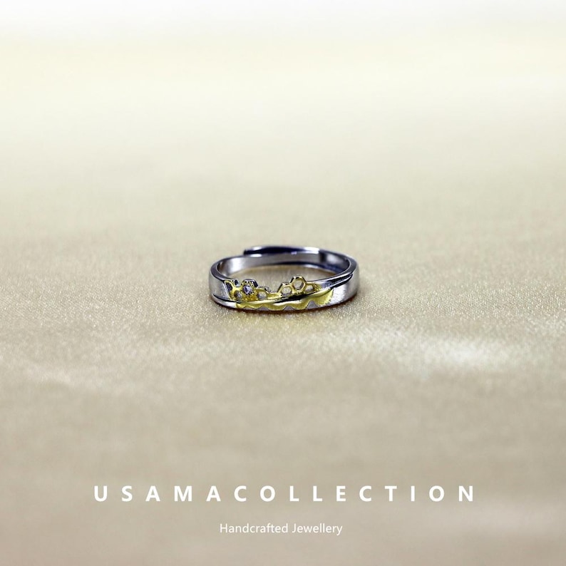 May include: A silver and gold-toned ring with a floral design. The ring has a band of silver with a gold-colored floral pattern across the top. The ring is on a neutral background. The text "USAMA COLLECTION" and "Handcrafted Jewellery" are at the bottom.