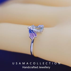 May include: A close-up of a silver ring with a butterfly design. The butterfly has purple and blue wings and a light-colored body. The ring is on a light-colored background. The text "USAMA COLLECTION Handcrafted Jewellery" is at the bottom.