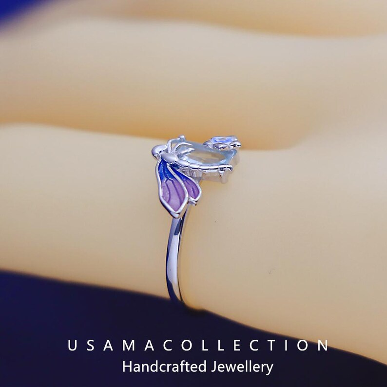 May include: A close-up of a silver ring with a butterfly design. The butterfly has purple and blue wings and a light-colored body. The ring is on a light-colored background. The text "USAMA COLLECTION Handcrafted Jewellery" is at the bottom.