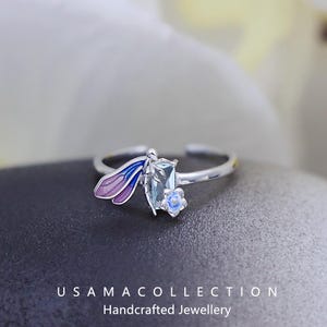 May include: A silver ring featuring a dragonfly design. The dragonfly has purple and blue wings, a rectangular blue-green gemstone body, and a small blue flower accent. The ring is open-ended and sits on a dark surface. The text "USAMA COLLECTION Handcrafted Jewellery" is at the bottom.