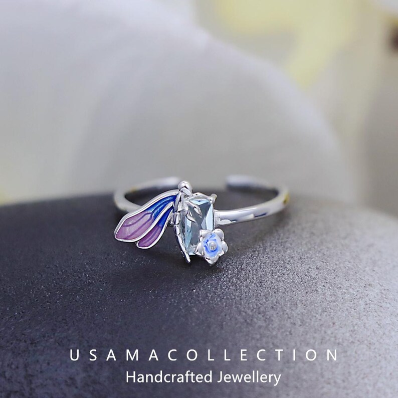 May include: A silver ring featuring a dragonfly design. The dragonfly has purple and blue wings, a rectangular blue-green gemstone body, and a small blue flower accent. The ring is open-ended and sits on a dark surface. The text "USAMA COLLECTION Handcrafted Jewellery" is at the bottom.