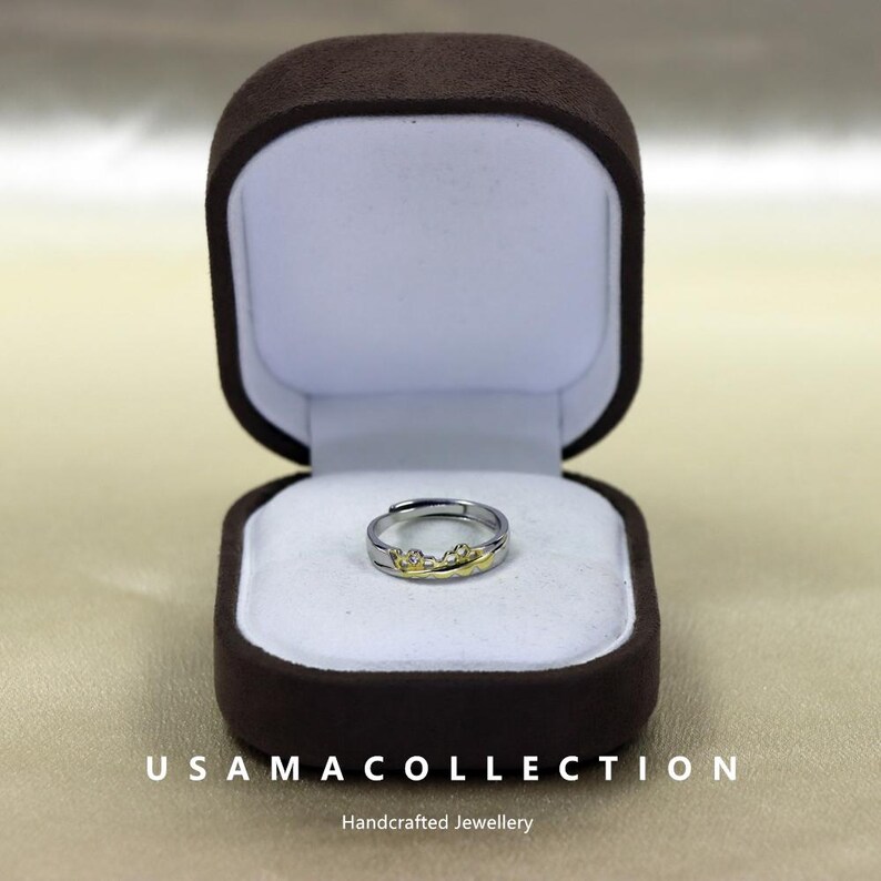 May include: A silver-toned ring with a gold-colored mountain range design, displayed inside a brown velvet ring box. The ring is open-ended and adjustable. The box is open, revealing the ring on a white satin interior.