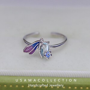 May include: A silver ring featuring a dragonfly design with purple and blue wings, a light blue gemstone, and a small blue flower. The ring is adjustable and sits on a light background. The text "USAMA COLLECTION Handcrafted Jewellery" is at the bottom.