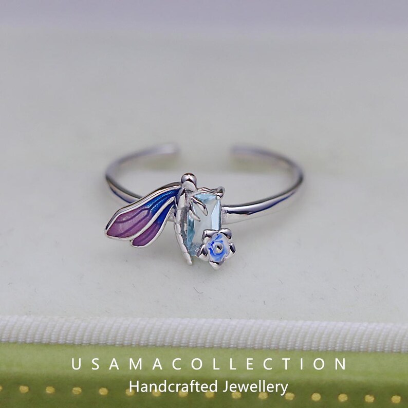 May include: A silver ring featuring a dragonfly design with purple and blue wings, a light blue gemstone, and a small blue flower. The ring is adjustable and sits on a light background. The text "USAMA COLLECTION Handcrafted Jewellery" is at the bottom.