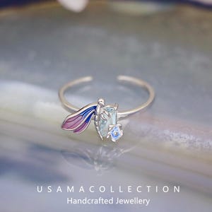 May include: A silver ring featuring a dragonfly design with blue and purple wings, a light blue rectangular gemstone, and a small blue flower. The ring is adjustable and has the text "USAMA COLLECTION Handcrafted Jewellery".