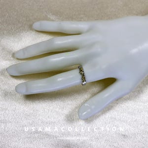 May include: A silver ring with gold accents is displayed on a mannequin hand. The ring features an intricate design with a repeating pattern. The ring is a delicate piece of jewelry.