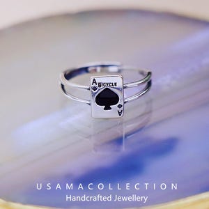 Può includere: Un anello d'argento con un motivo a carta da gioco. La carta è l'Asso di Picche di un mazzo Bicycle, con il simbolo di picche nero. L'anello ha un design a doppia fascia ed è ambientato su uno sfondo sfocato e colorato.