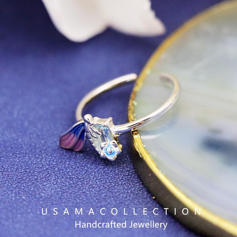 May include: A silver ring with a floral design. The ring features a light blue gemstone and a small blue and pink flower. The ring is on a blue surface with the text "USAMA COLLECTION Handcrafted Jewellery".