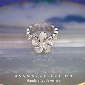 May include: A silver-toned adjustable ring featuring a flower design. The flower's petals are encrusted with small, sparkling stones, and a larger stone is at the center. The ring's band is adorned with heart-shaped cutouts. The text "USAMA COLLECTION Handcrafted Jewellery" is at the bottom.