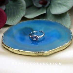 May include: A silver adjustable ring with a blue gemstone, set on a blue and gold decorative plate. The ring has a textured band and a central oval-shaped stone. The plate has a gold-colored edge and a polished surface.