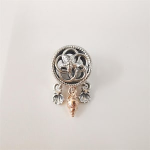 Könnte beinhalten: Ein silberner Charm mit einem Seestern und einem kreisförmigen Design, akzentuiert mit einem goldfarbenen Muschelanhänger und zwei silbernen Muschel-Charms. Der Charm ist vor einem weißen Hintergrund platziert.