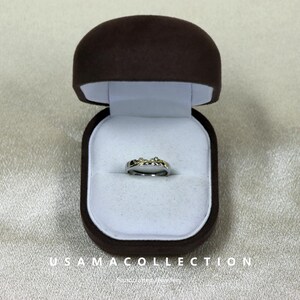 May include: A silver ring with small, sparkling stones is displayed inside a brown velvet ring box with a white interior. The ring box is open, showcasing the ring's design. The text "USAMA COLLECTION" is visible at the bottom of the image.