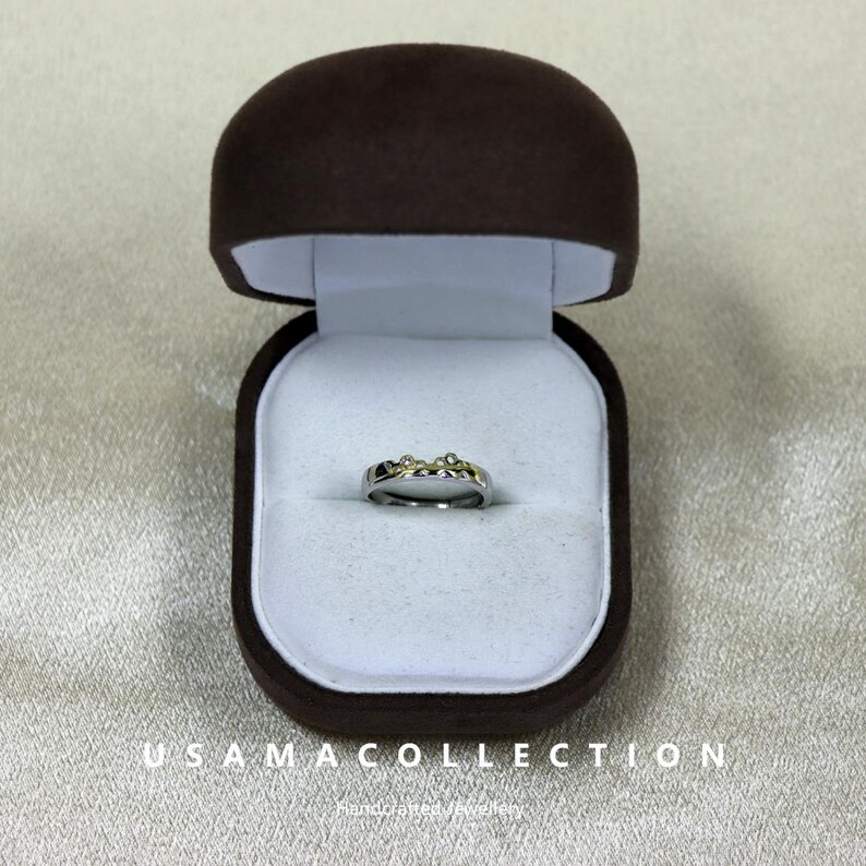 May include: A silver ring with small, sparkling stones is displayed inside a brown velvet ring box with a white interior. The ring box is open, showcasing the ring's design. The text "USAMA COLLECTION" is visible at the bottom of the image.