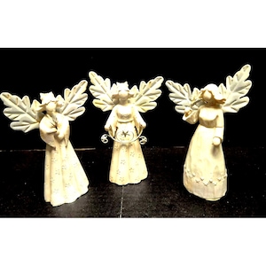 May include: Three off-white angel figurines with detailed wings and flowing gowns. Each angel has a unique pose, with one holding a star-shaped object. The figurines are likely made of a ceramic or resin material.