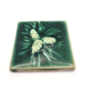May include: Square ceramic tile with a green glaze and a raised floral design. The tile has a glossy finish and a crackle effect. The floral design is in a lighter shade of green, creating a contrast against the darker background.