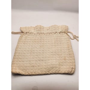 May include: A cream-colored drawstring pouch crafted from crocheted yarn. The bag features a textured, repeating stitch pattern throughout the body and a scalloped edge along the top opening. Two matching crocheted cords are threaded through the top.