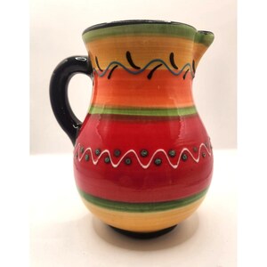 May include: A colorful ceramic pitcher with a black handle and spout. The pitcher features horizontal bands of orange, red, yellow, and green, with decorative wavy lines and dots. The pitcher is approximately 7 inches tall.