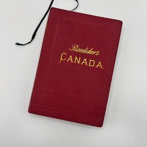 May include: A vintage red Baedeker's Canada travel guide with gold lettering and a dark green ribbon bookmark. The book's cover features the title "Baedeker's CANADA."