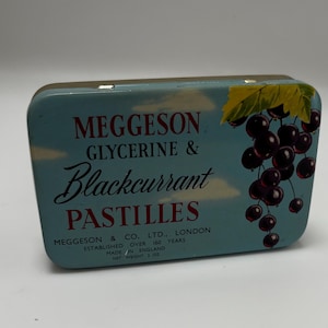 May include: Vintage Meggeson Glycerine & Blackcurrant Pastilles tin. The rectangular tin has a light blue background with the product name in red and black script. A cluster of blackcurrants and leaves are on the right side. The tin is marked "Made in England".