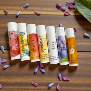May include: Seven lip balm tubes with white caps and colorful labels. Flavors include Watermelon, Osmanthus, Mango, Pineapple, Rose, Grape, and Orange. The tubes are arranged on a wooden surface with scattered purple and blue decorative elements.