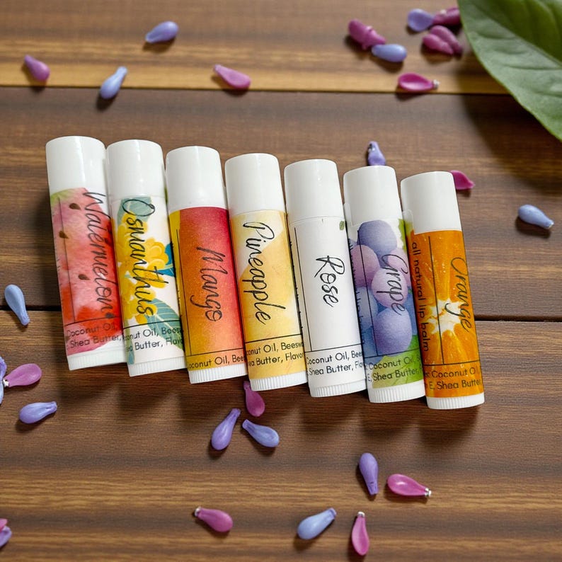 May include: Seven lip balm tubes with white caps and colorful labels. Flavors include Watermelon, Osmanthus, Mango, Pineapple, Rose, Grape, and Orange. The tubes are arranged on a wooden surface with scattered purple and blue decorative elements.